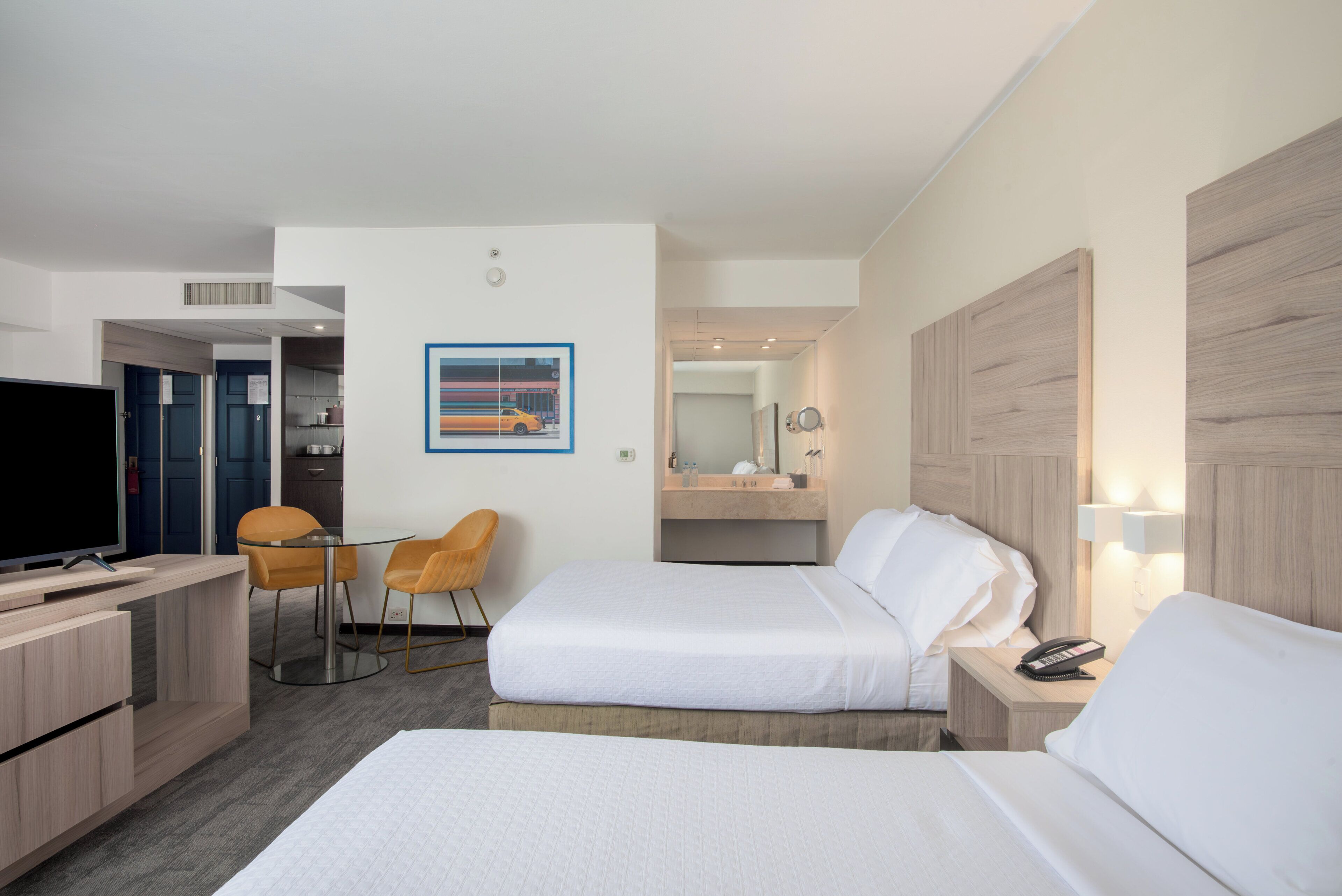 Habitación de hotel con dos camas, televisión, escritorio con silla, baño y un pequeño comedor.