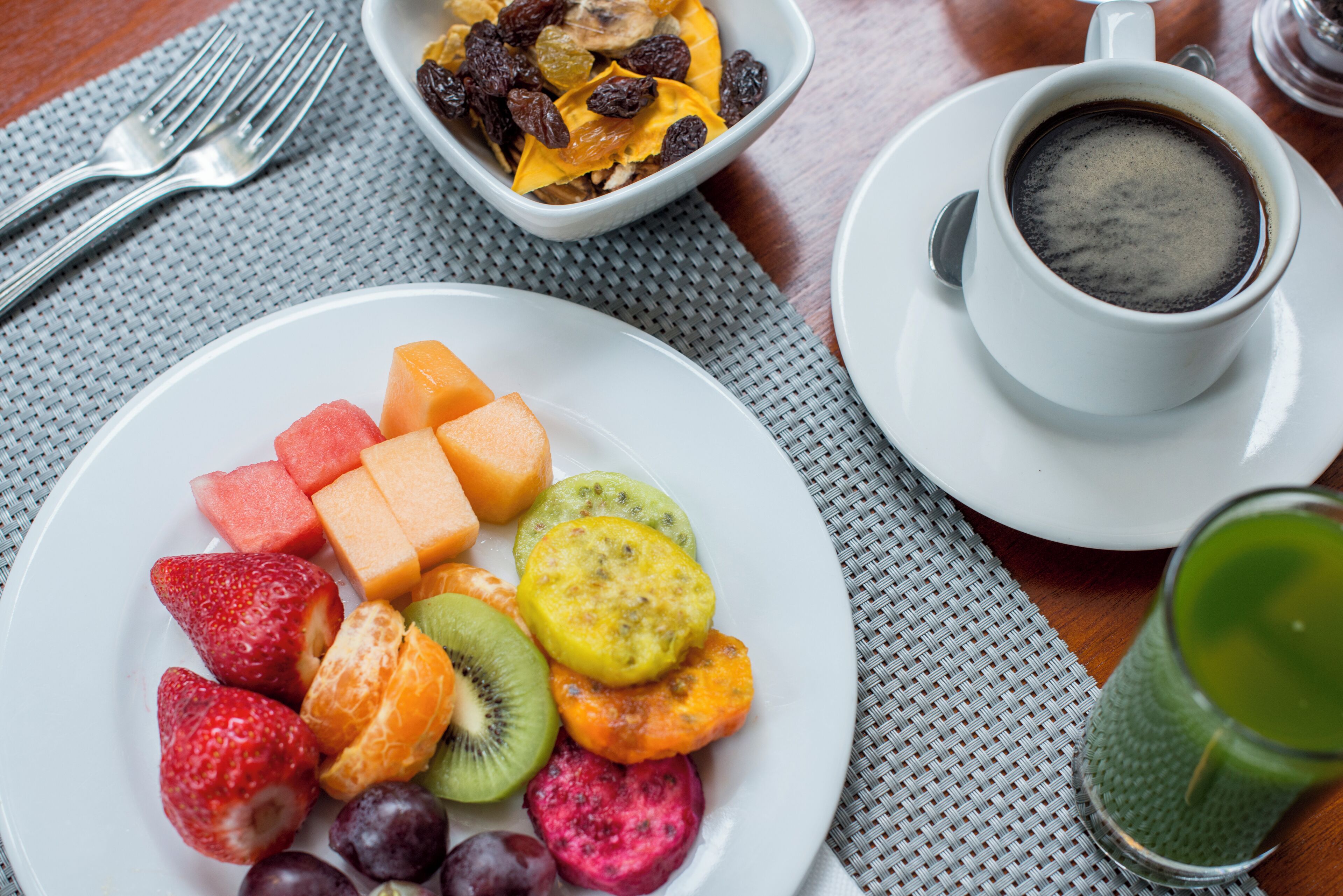 A breakfast setting with a plate of assorted fruits, a bowl of cereal with raisins, a cup of coffee, and a glass of green juice.