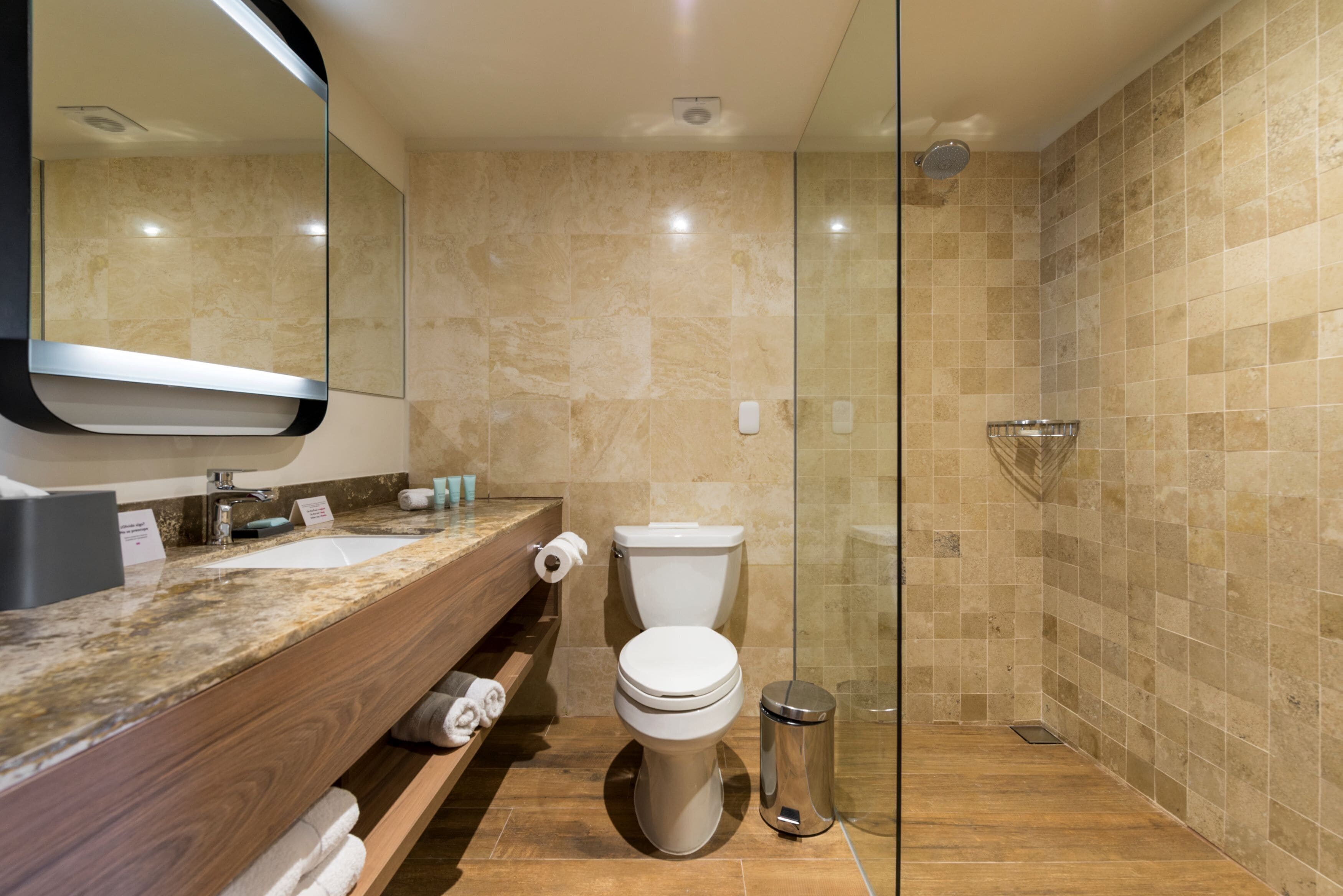 A modern bathroom with a marble countertop, a toilet, a glass shower enclosure, and a trash can.