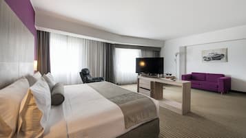 A hotel room with a large bed, a desk with a TV, a purple sofa, and a car-themed artwork on the wall.