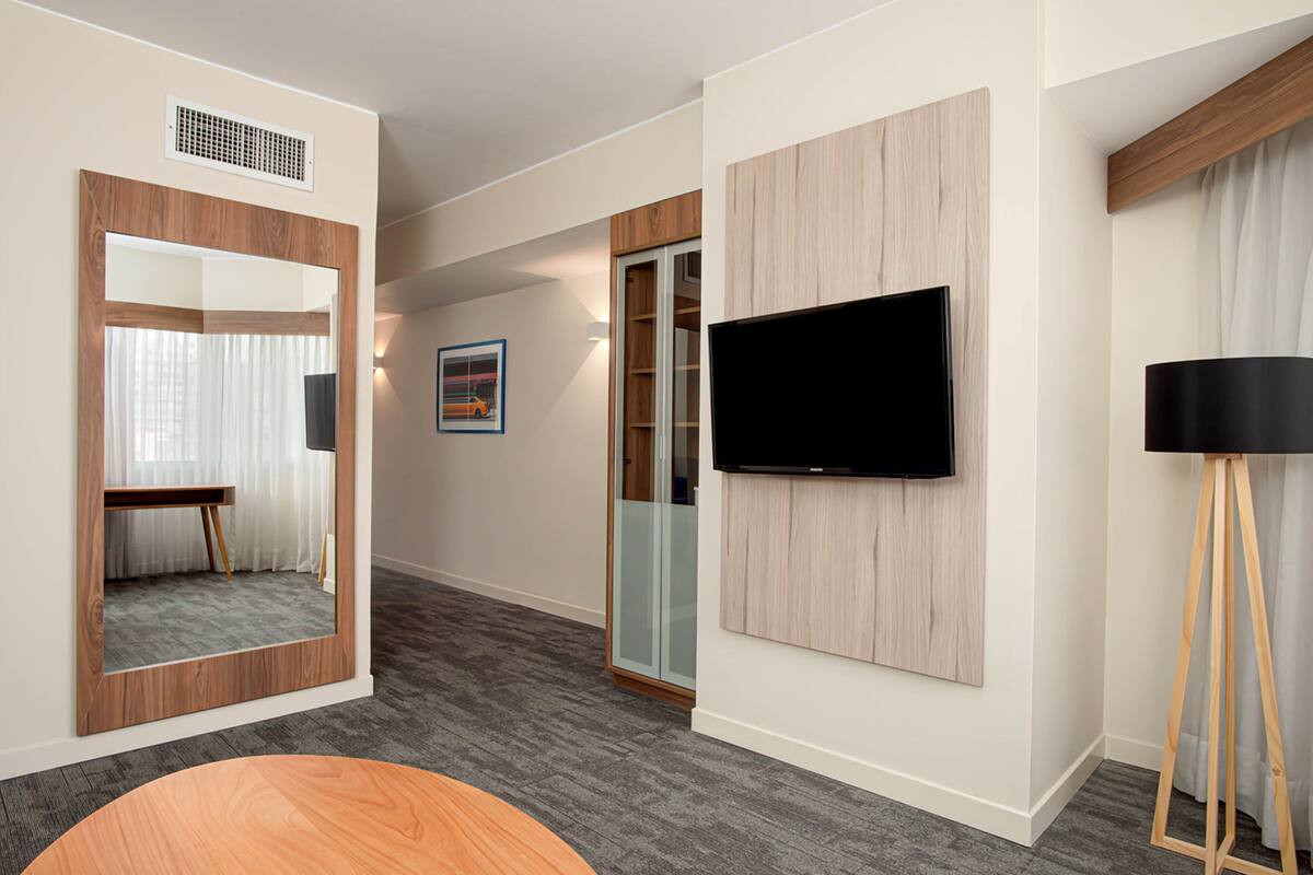 A modern hotel room with a large mirror, a flat-screen TV mounted on the wall, a wooden floor, and a glass door leading to a balcony.