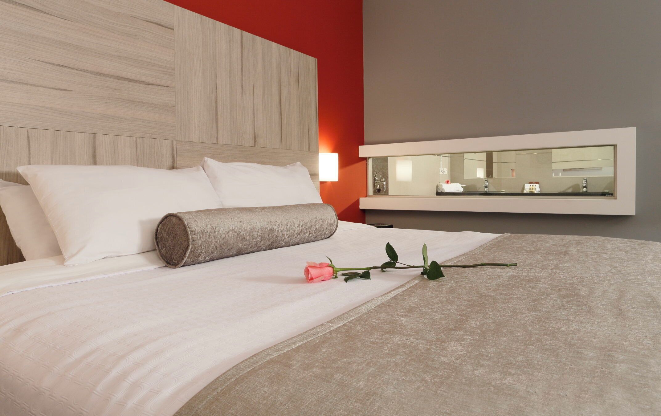 Ein Hotelzimmer mit Bett, Nachttischlampe, Spiegel und einer dekorativen Rose auf dem Bett.