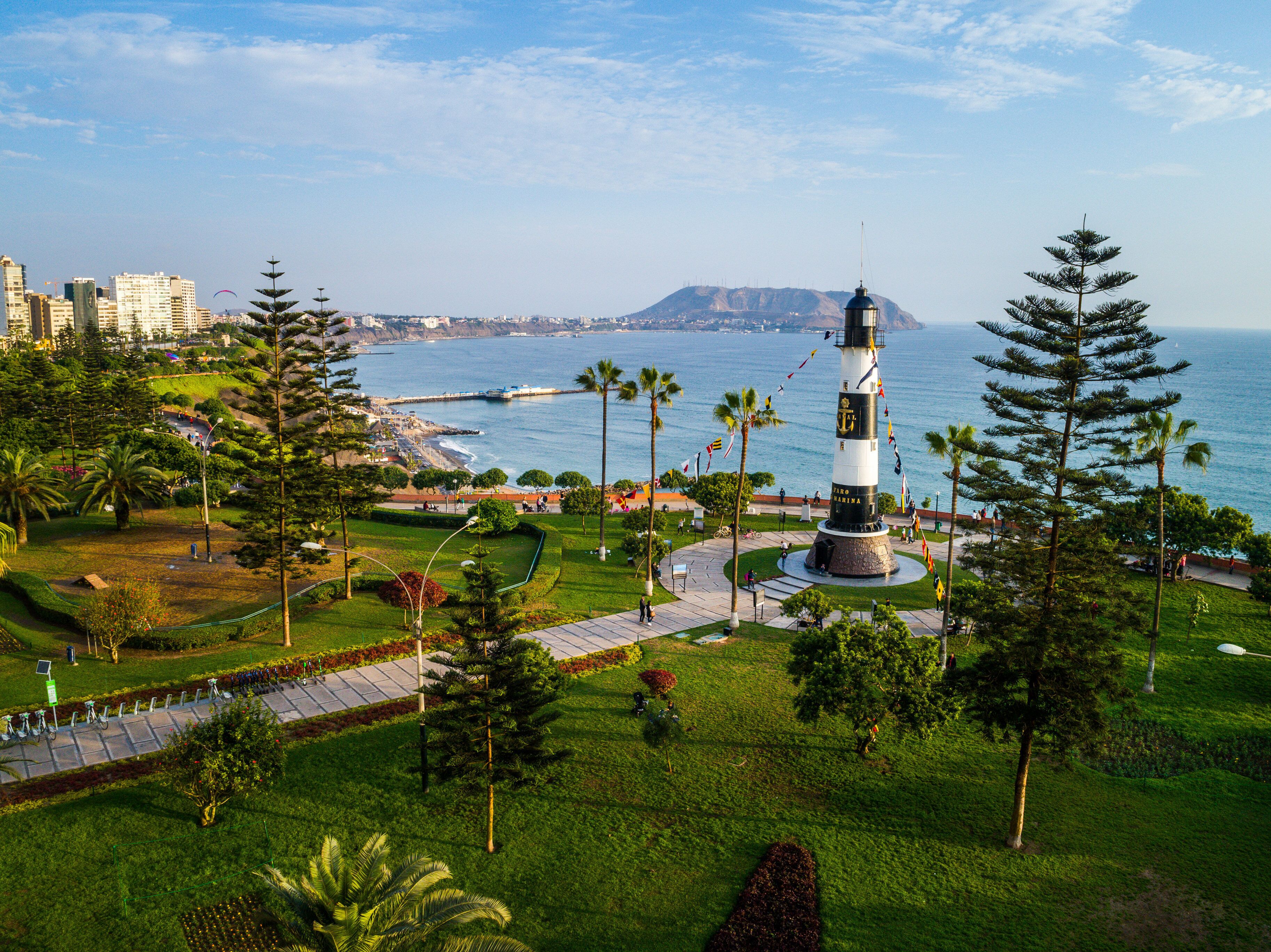 Un faro rodeado por un parque con senderos y palmeras, con vistas a una ciudad costera.