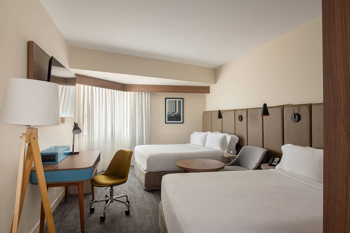 A hotel room with two beds, a desk with a lamp, a chair, a small table, and a painting on the wall.