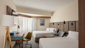 A hotel room with two beds, a desk with a lamp, a chair, a small table, and a painting on the wall.