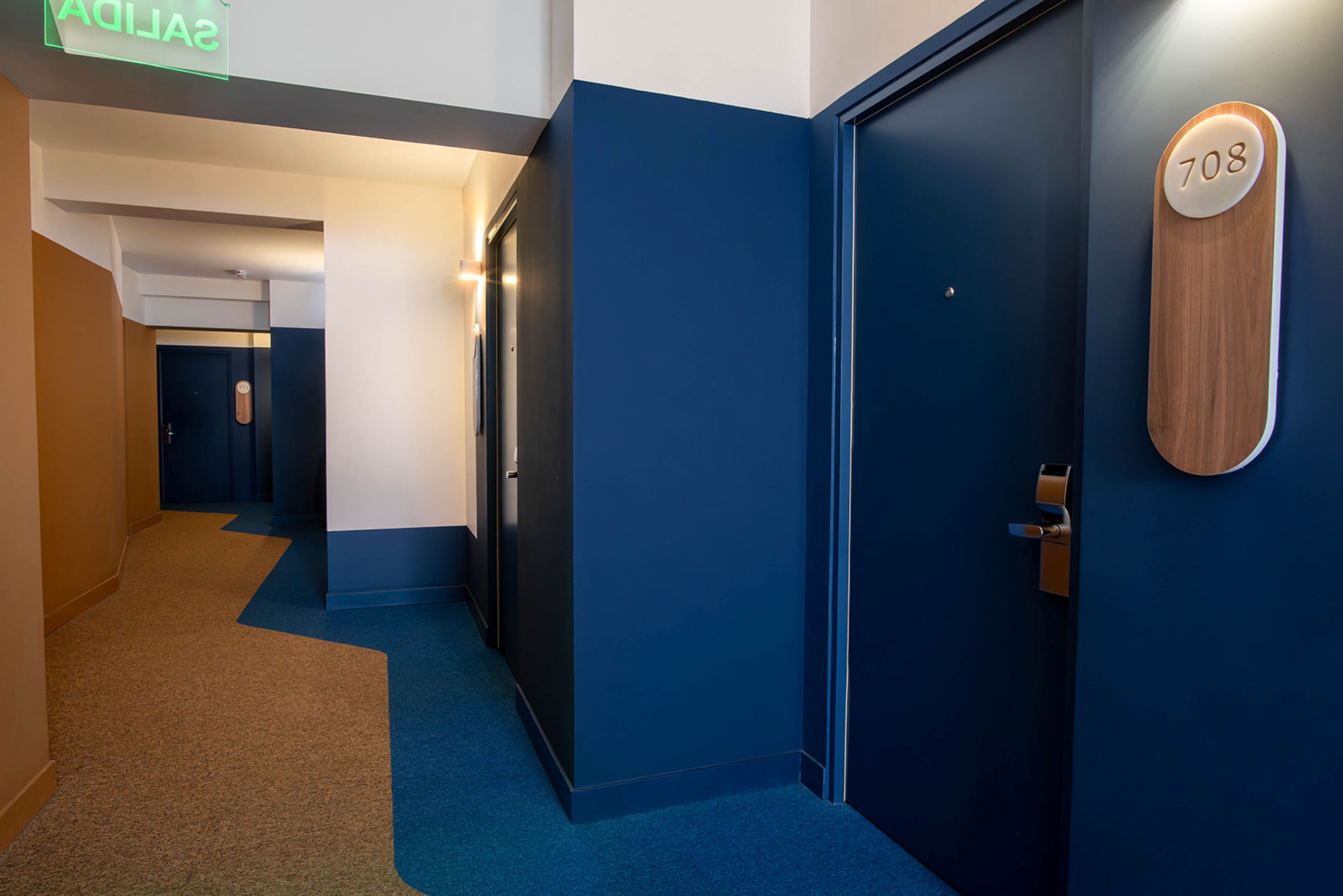 A modern hotel corridor with blue walls and carpeted floors, featuring a wooden door with the number 708.