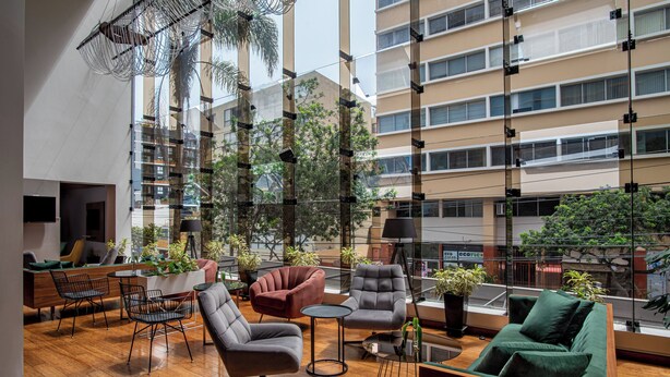 A spacious indoor area with modern furniture, including sofas and chairs, and a large window offering a view of the surrounding buildings. - Crowne Plaza Lima - Miraflores by IHG (Lima)