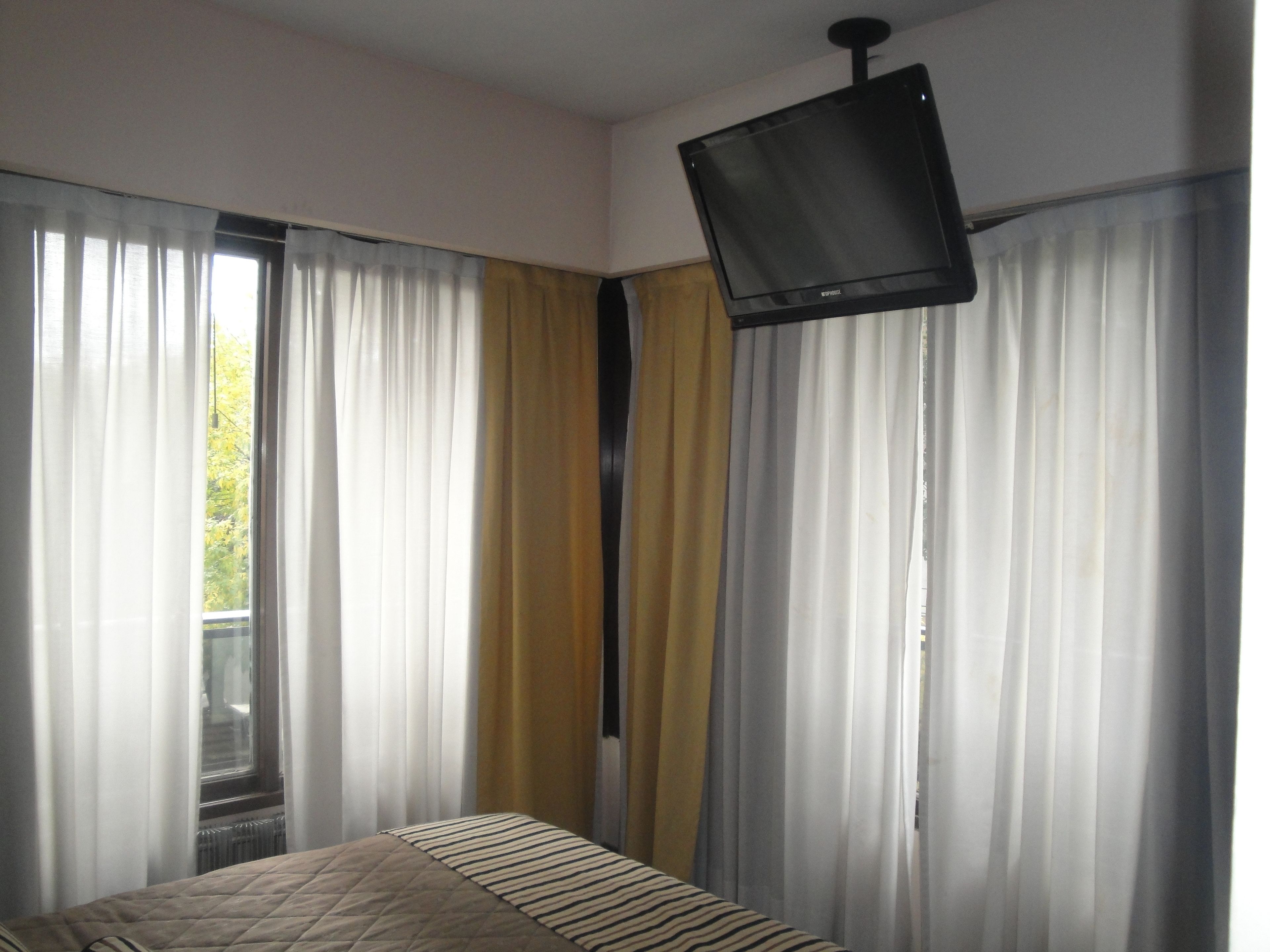 A bedroom with a bed, a window with white and yellow curtains, and a mounted television.