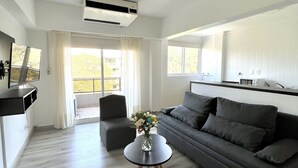 A modern living room with a grey sofa, a round coffee table with a vase, and a grey ottoman. There is a kitchen area with a white countertop and a built-in oven. - Golf Tower Suites & Apartments (Buenos Aires)