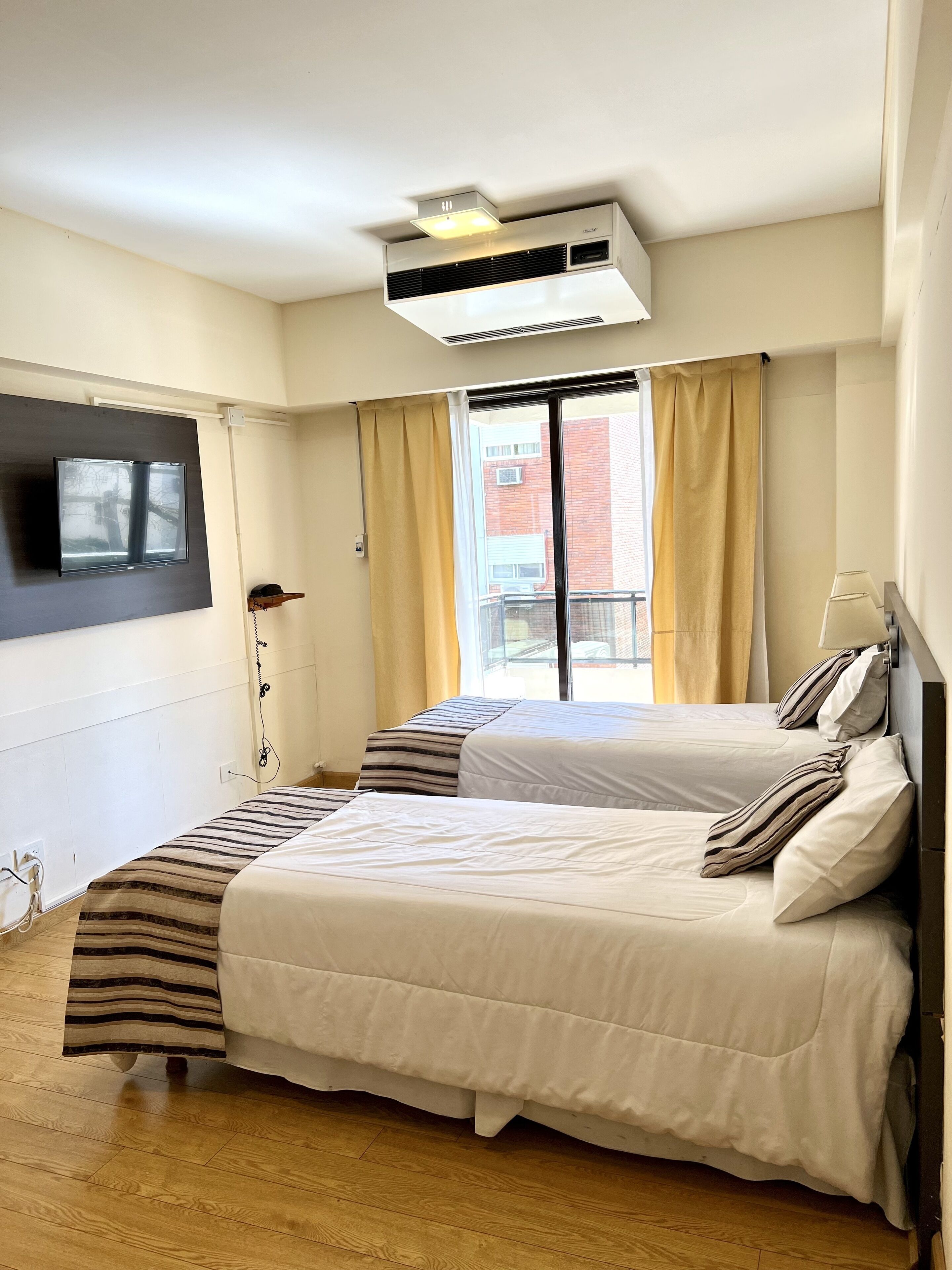 A hotel room with two beds, a flat-screen TV mounted on the wall, an air conditioning unit, and a window with curtains.