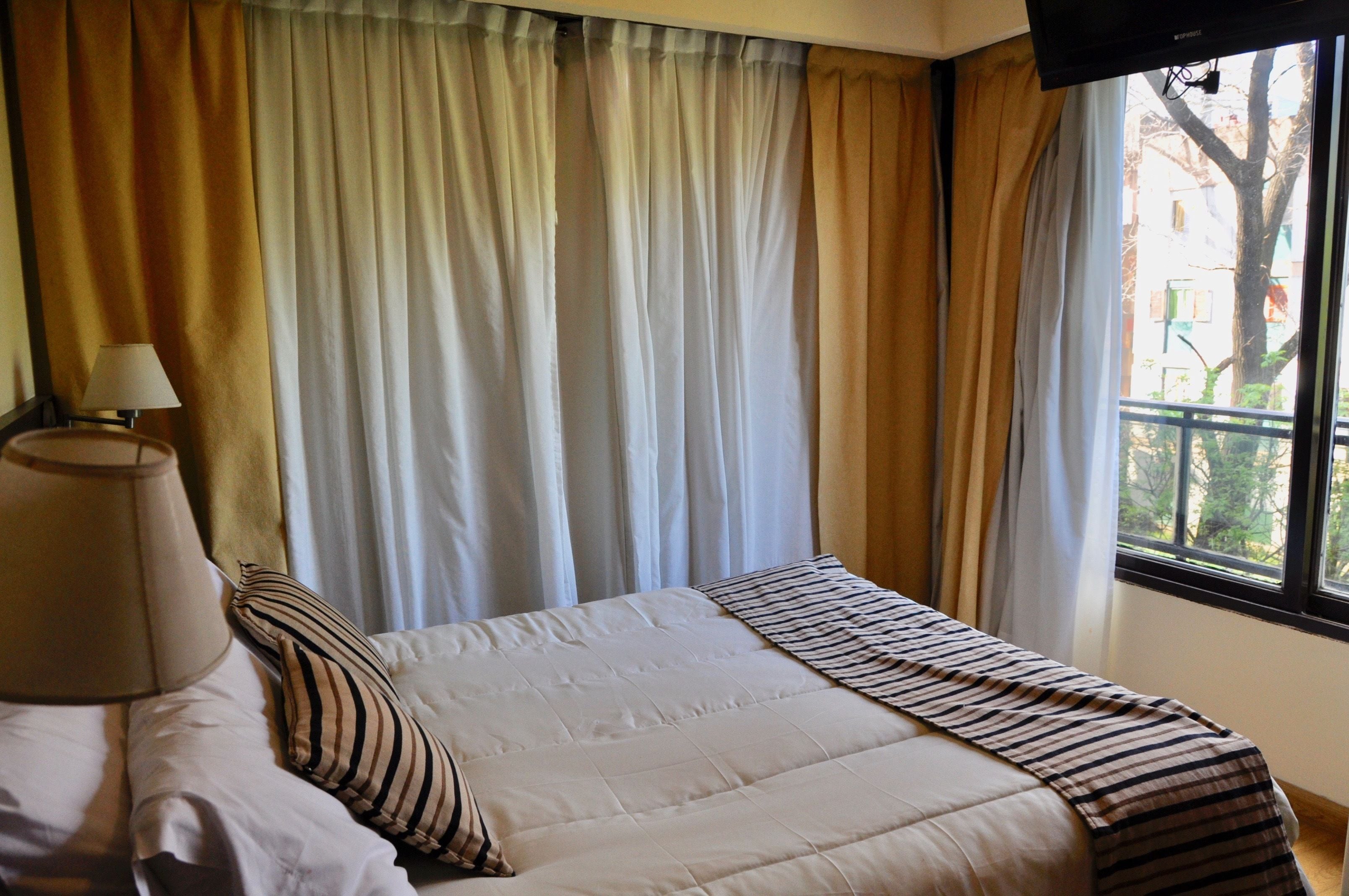 A hotel room with a bed, curtains, a lamp, and a window with a view of buildings and trees.