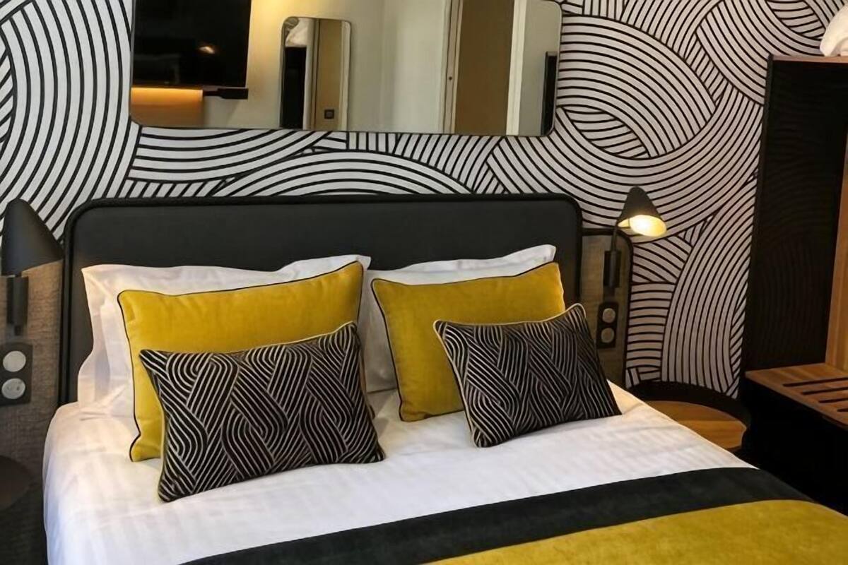 A bed with white and yellow bedding, two patterned pillows, a black headboard, and a mirror above the bed.