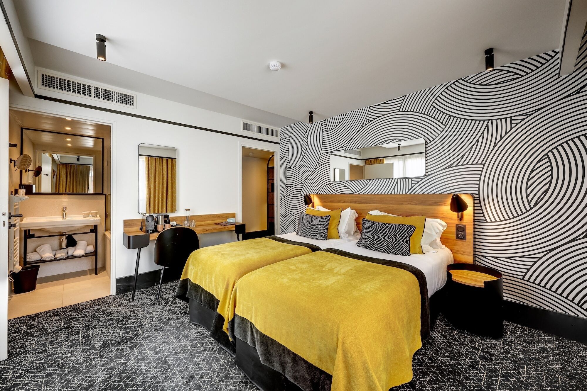 Una habitación de hotel con una cama, un tocador con espejo, un escritorio y un papel tapiz estampado.