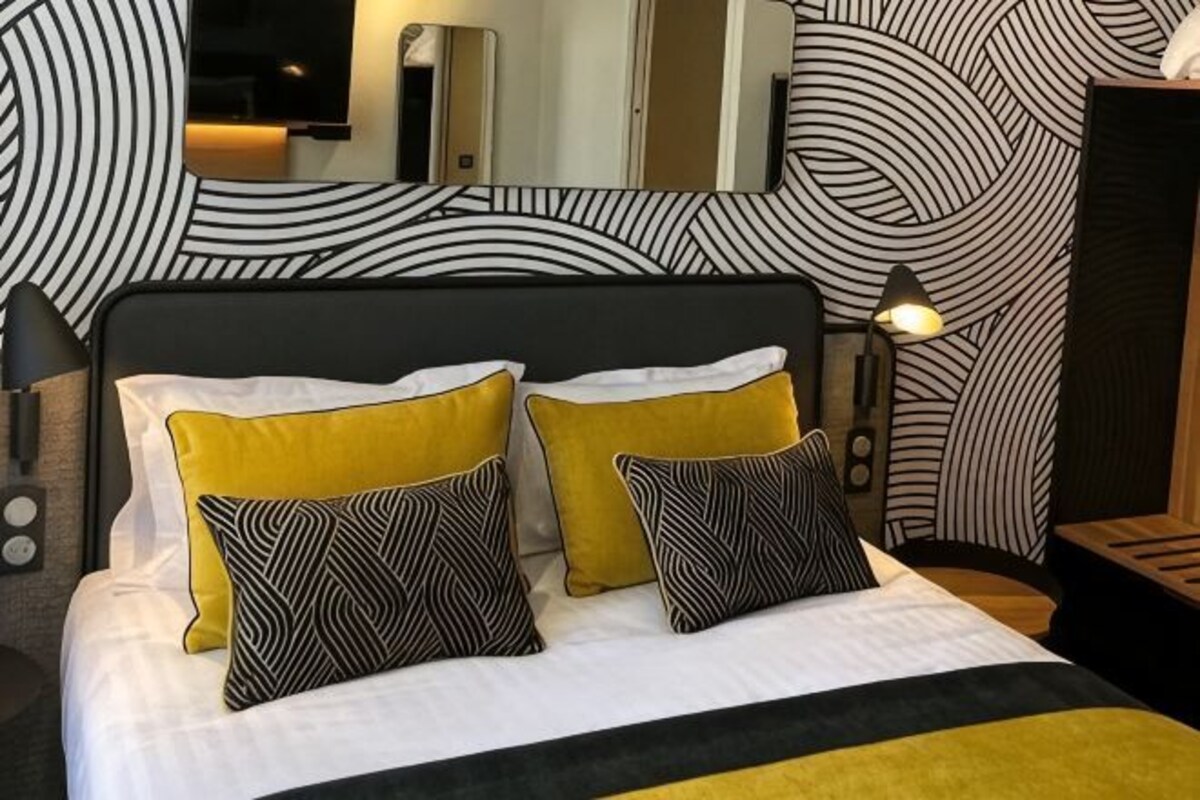 Una cama con ropa de cama blanca y amarilla, dos almohadas estampadas, un cabecero negro y un espejo sobre la cama.