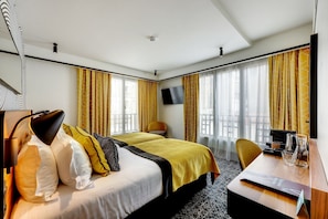 Quarto de hotel com duas camas, uma escrivaninha, uma cadeira, uma televisão e amplas janelas com cortinas.