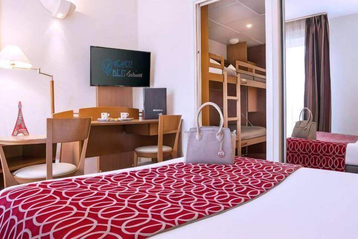 A hotel room with a bed, a desk with a chair, a television, a bunk bed, and a handbag on the bed.