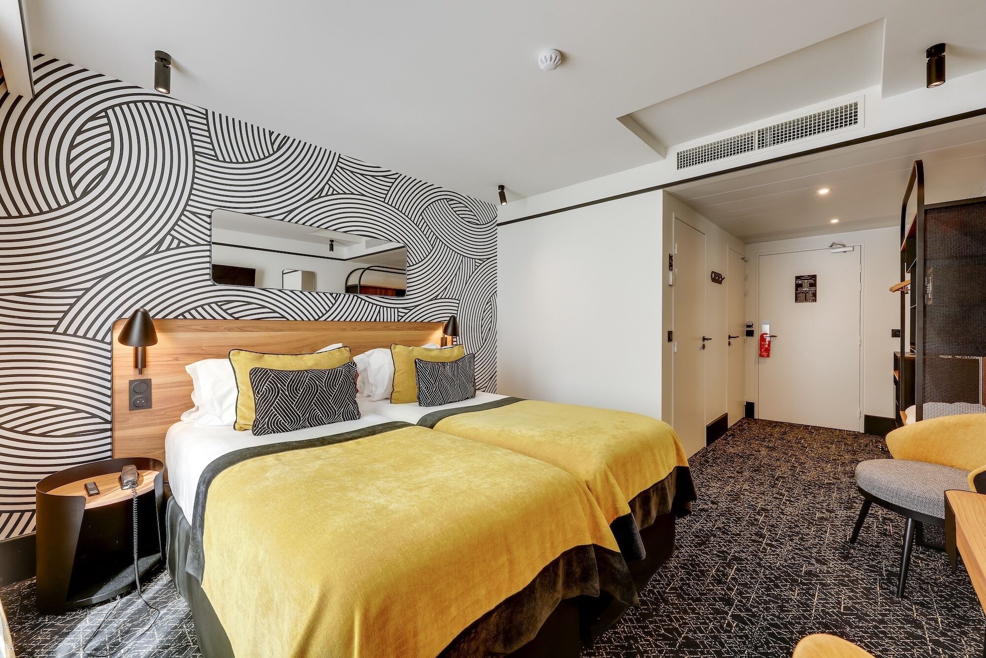 Una habitación de hotel con cama, mesitas de noche, una silla, un espejo y una pared decorada.