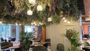 An indoor seating area with round tables and chairs, surrounded by potted plants and hanging lights. - Hôtel Vacances Bleues Villa Modigliani (Paris)
