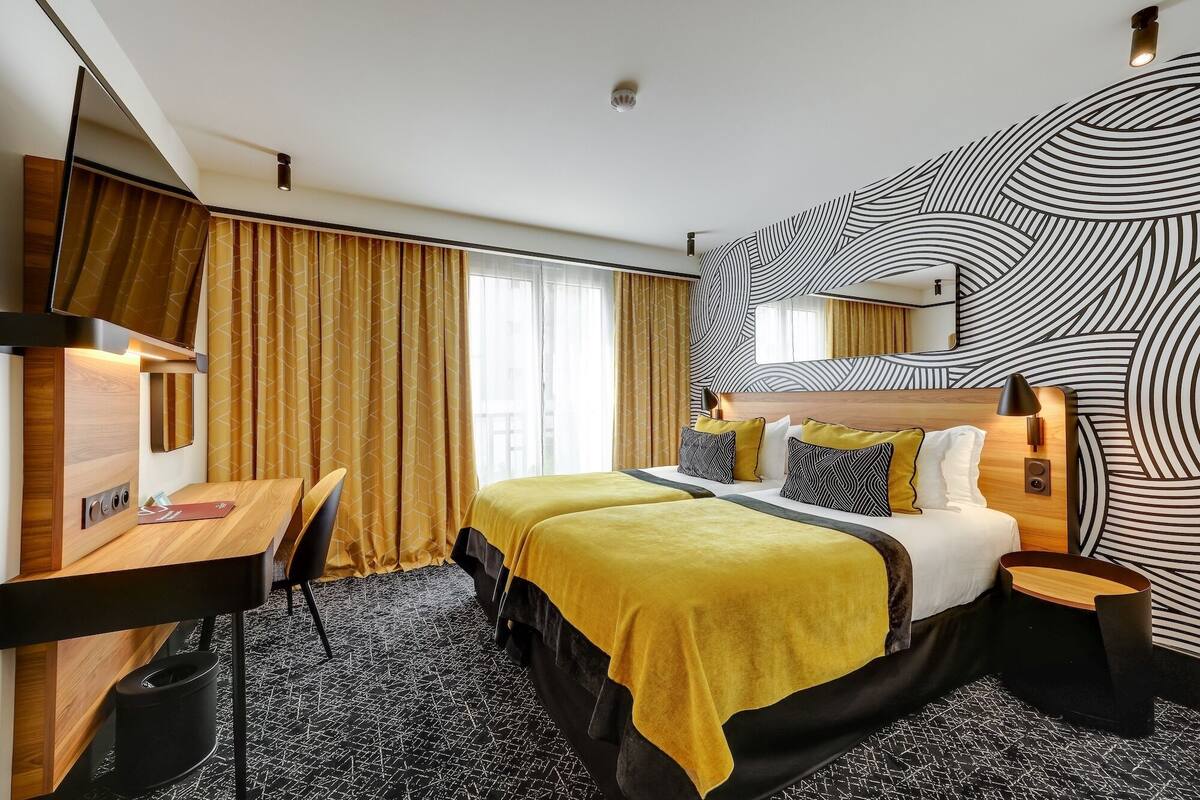 A hotel room with a bed, desk, and chair, a large mirror, and a patterned wall.