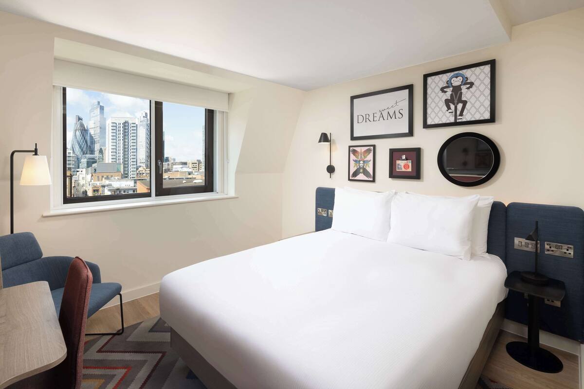 A hotel room with a large bed, a desk, a chair, a window with a city view, and wall art.