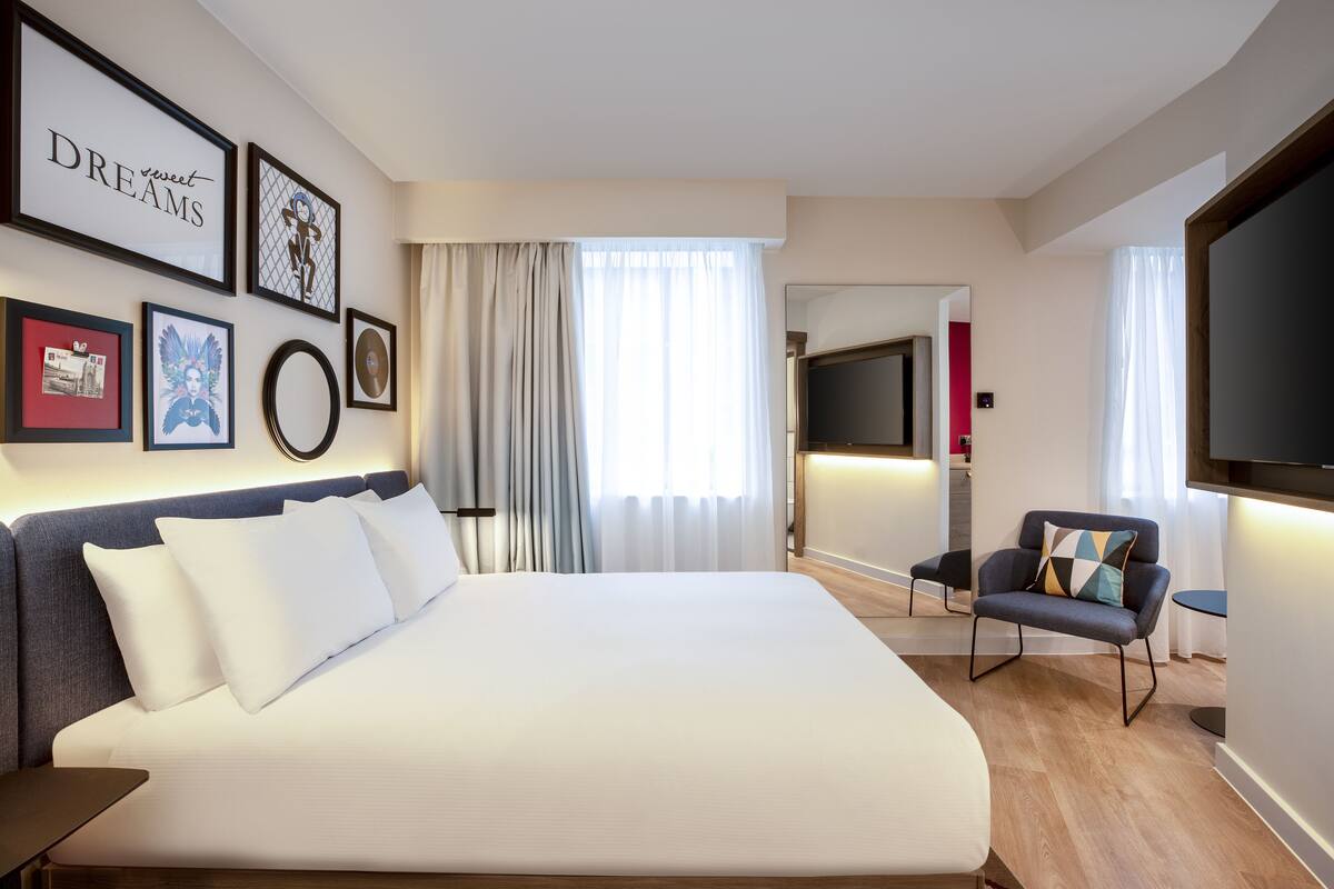 A modern hotel room with a large bed, a TV, a seating area, and artwork on the walls.