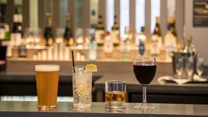 A bar area with three glasses of different beverages, a bottle of wine, and a bar counter with various bottles in the background. - Hampton by Hilton London City (London)
