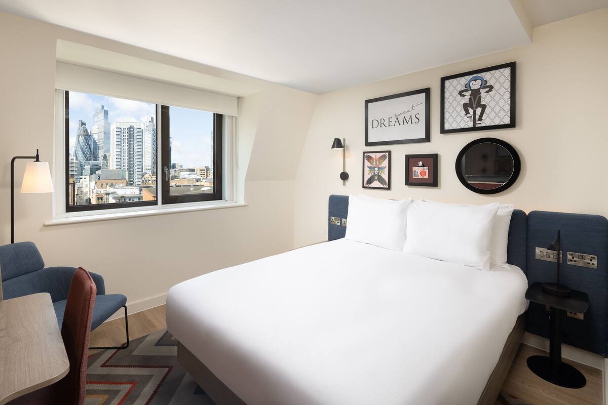 A hotel room with a large bed, a desk, a chair, a window with a city view, and several framed pictures on the wall.