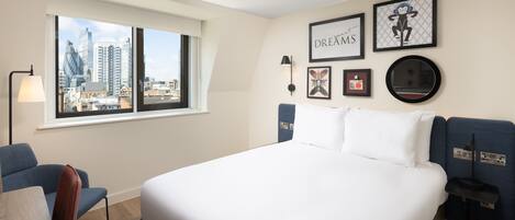 A hotel room with a large bed, a desk, a chair, a window with a city view, and several framed pictures on the wall.