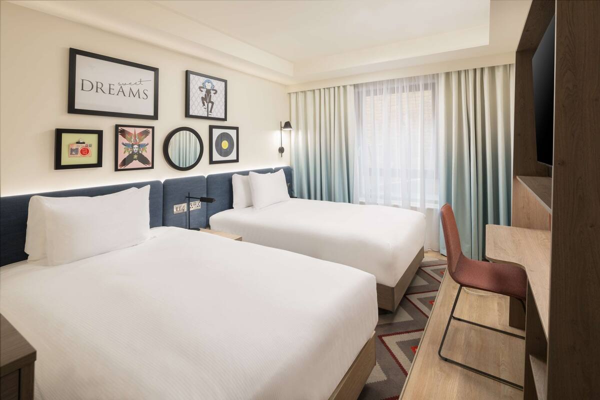A hotel room with two beds, a desk, and a chair. There are framed pictures on the wall, a window with curtains, and a light fixture.