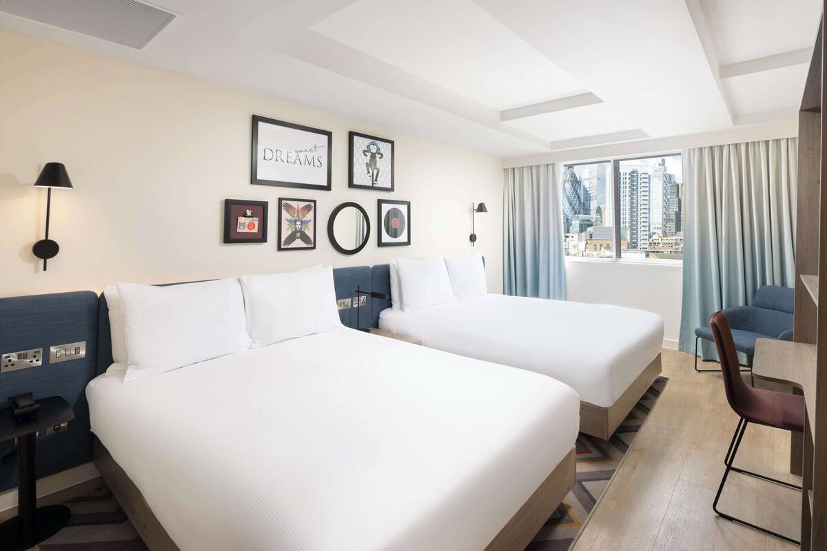 A hotel room with two beds, a desk, a chair, and a view of the city.