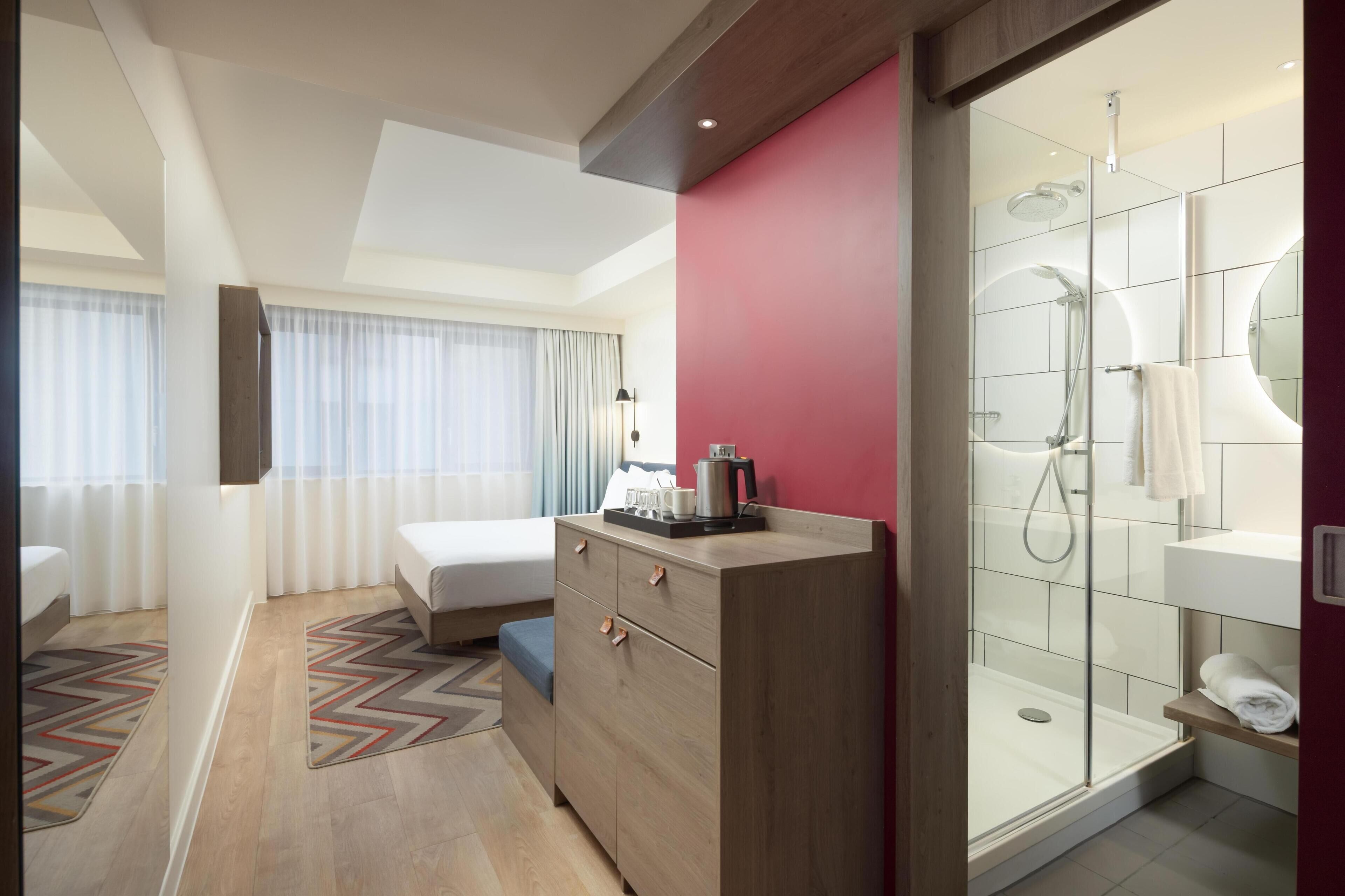 Ett modernt hotellrum med en säng, en byrå i trä, en duschkabin med glasväggar och utsikt över staden genom fönstret.