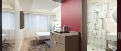 Ett modernt hotellrum med en säng, en byrå i trä, en duschkabin med glasväggar och utsikt över staden genom fönstret.