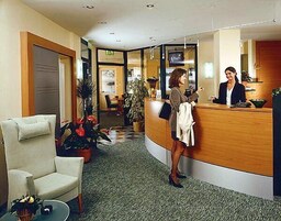 Zona reception di un hotel con una recezionista, una scrivania della reception, un'area salotto con un divano e una sala da pranzo sullo sfondo.