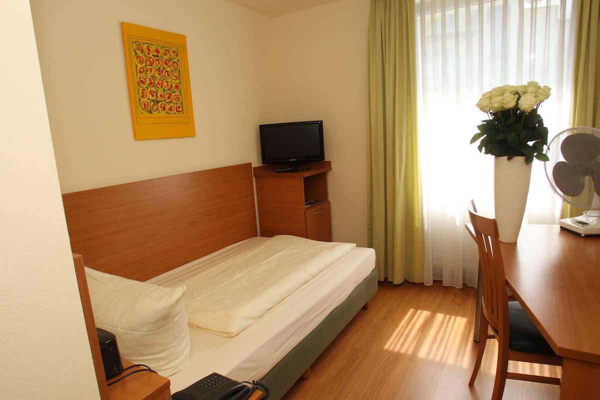 Une chambre d’hôtel avec un lit simple, une télévision, un bureau avec une chaise, un ventilateur et une fenêtre avec des rideaux.