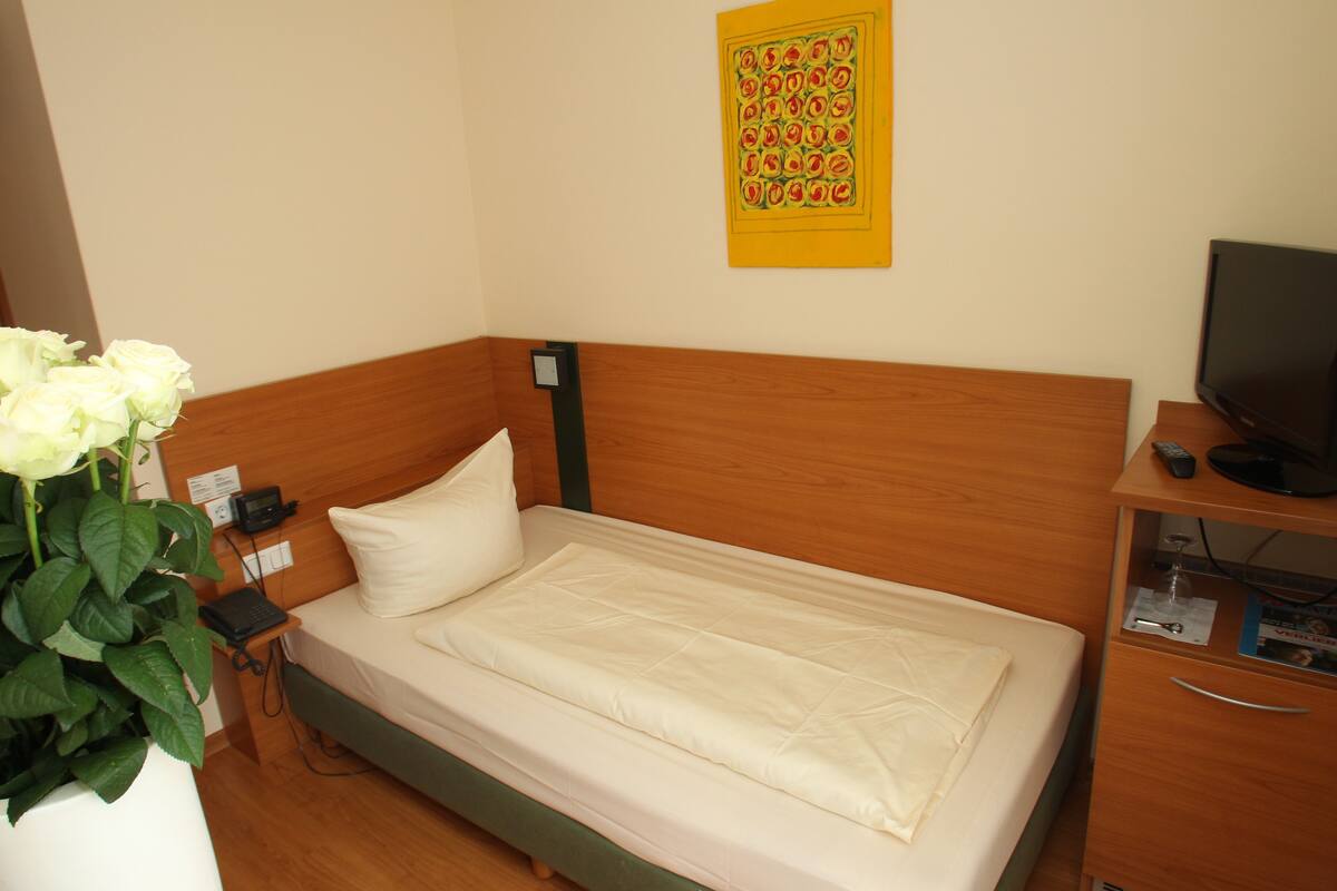 Un lit simple avec une tête de lit en bois, une table de chevet avec un téléphone et une télécommande, une petite armoire contenant un téléviseur, et une œuvre d’art accrochée au mur.