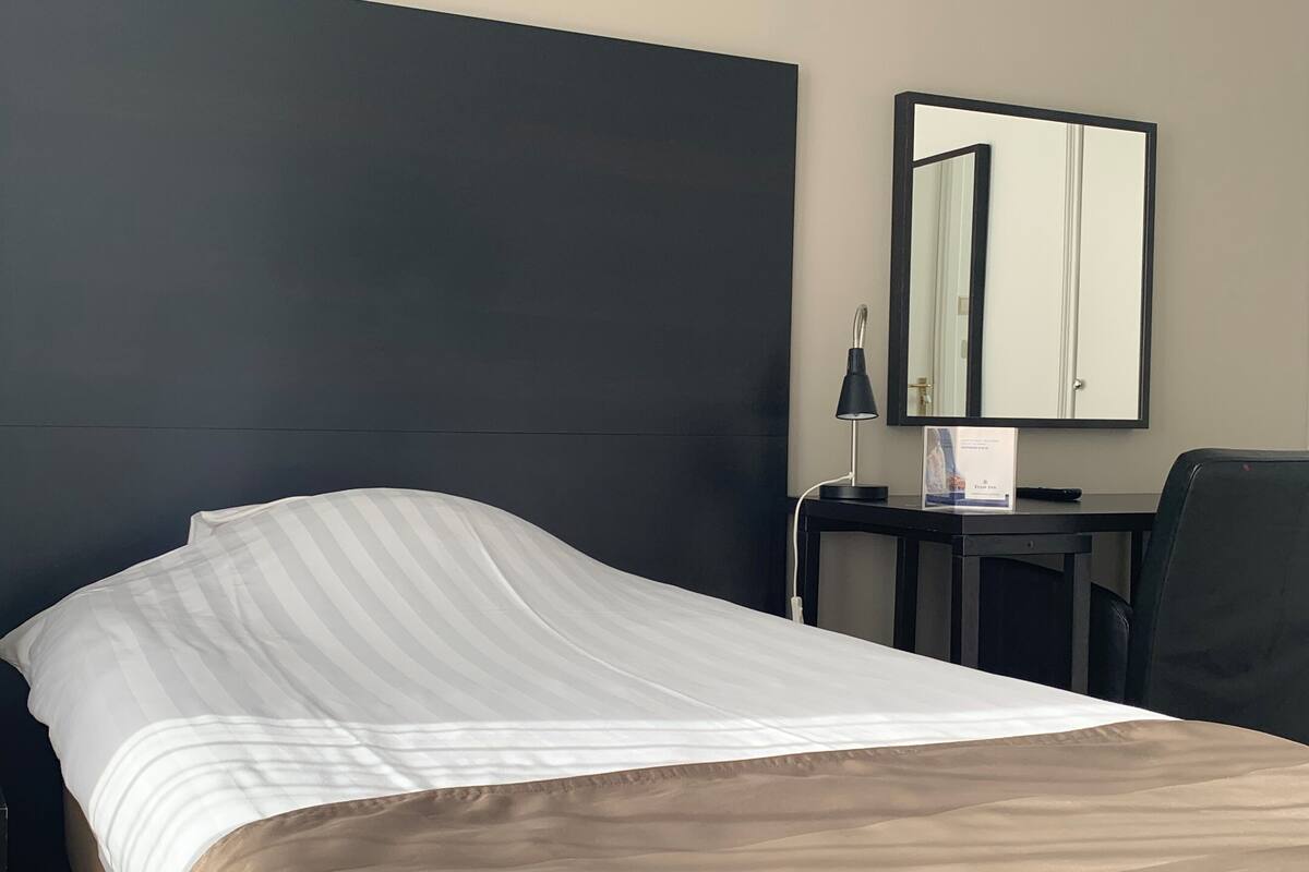 A hotel room with a bed, a desk, a chair, a lamp, and a mirror.
