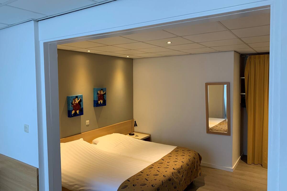 A hotel room with a bed, a nightstand, a mirror, and two paintings on the wall.