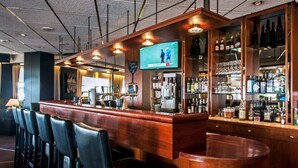 A bar area with a wooden counter, bar stools, a television screen, and shelves with bottles and glasses.