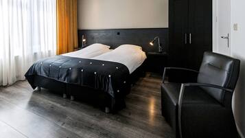 Quarto de hotel moderno com uma cama grande, uma mesa de cabeceira com abajur, uma poltrona de couro preta e um guarda-roupa.