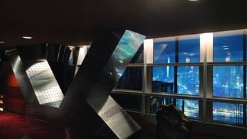 A modern interior with a large metallic sculpture, a view of a cityscape through large windows, and a polished wooden floor.