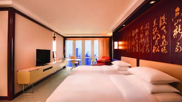 A hotel room with a large bed, a desk with a television, and a city view through the windows.
