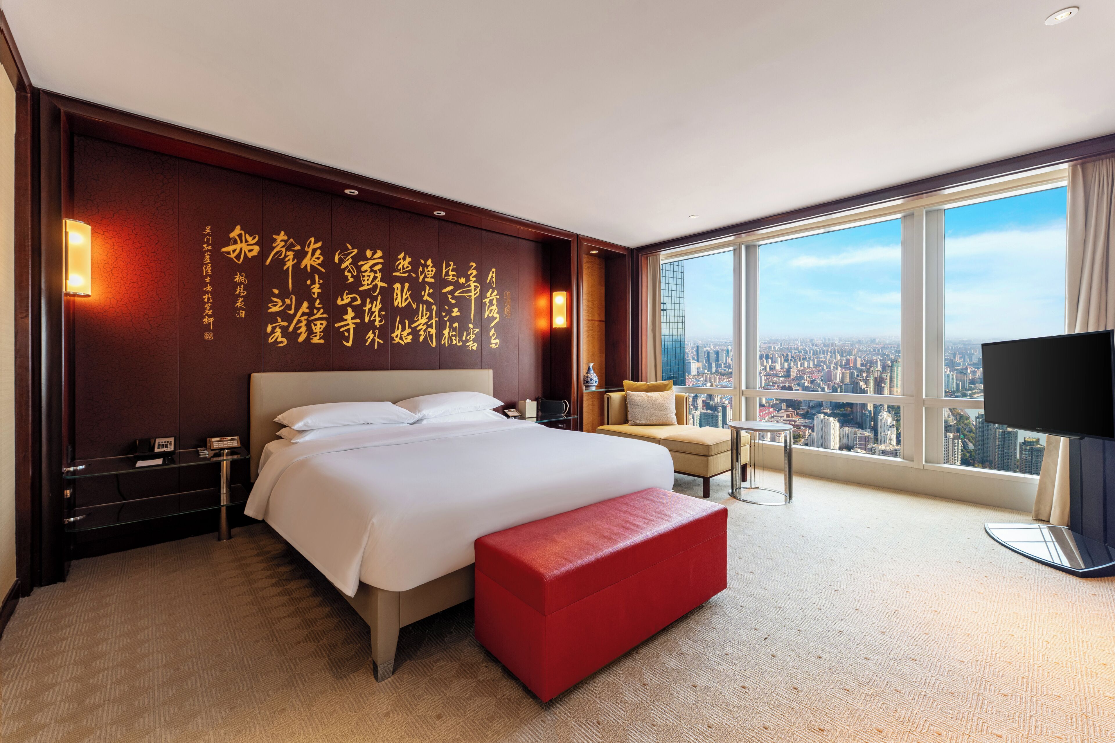 Ein Hotelzimmer mit einem großen Bett, einer roten Bank, einem Fernseher und Blick auf die Stadt.