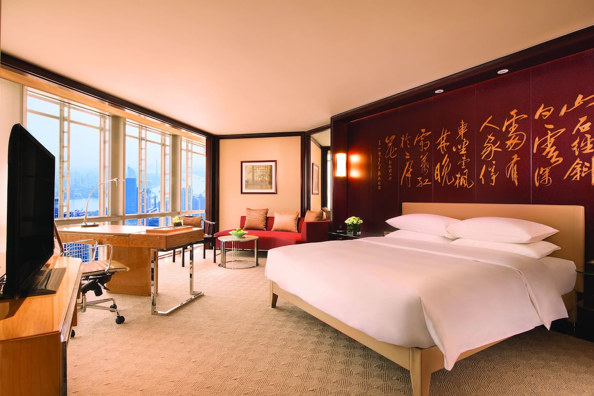 A hotel room with a large bed, a desk with a computer, a red sofa, and a large window offering a city view.