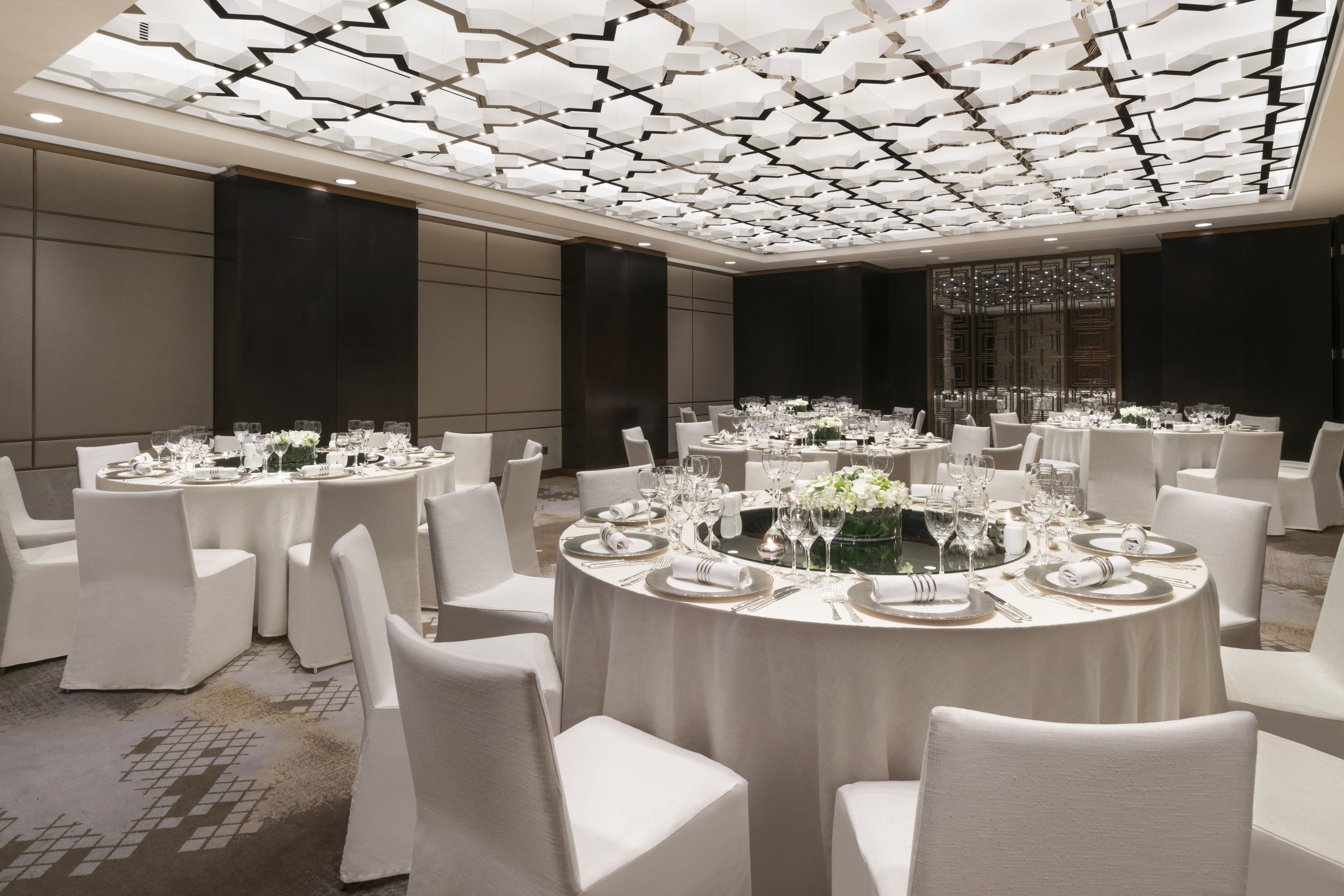 Un salón de banquetes con mesas redondas dispuestas para comer, sillas blancas y un diseño moderno en el techo.