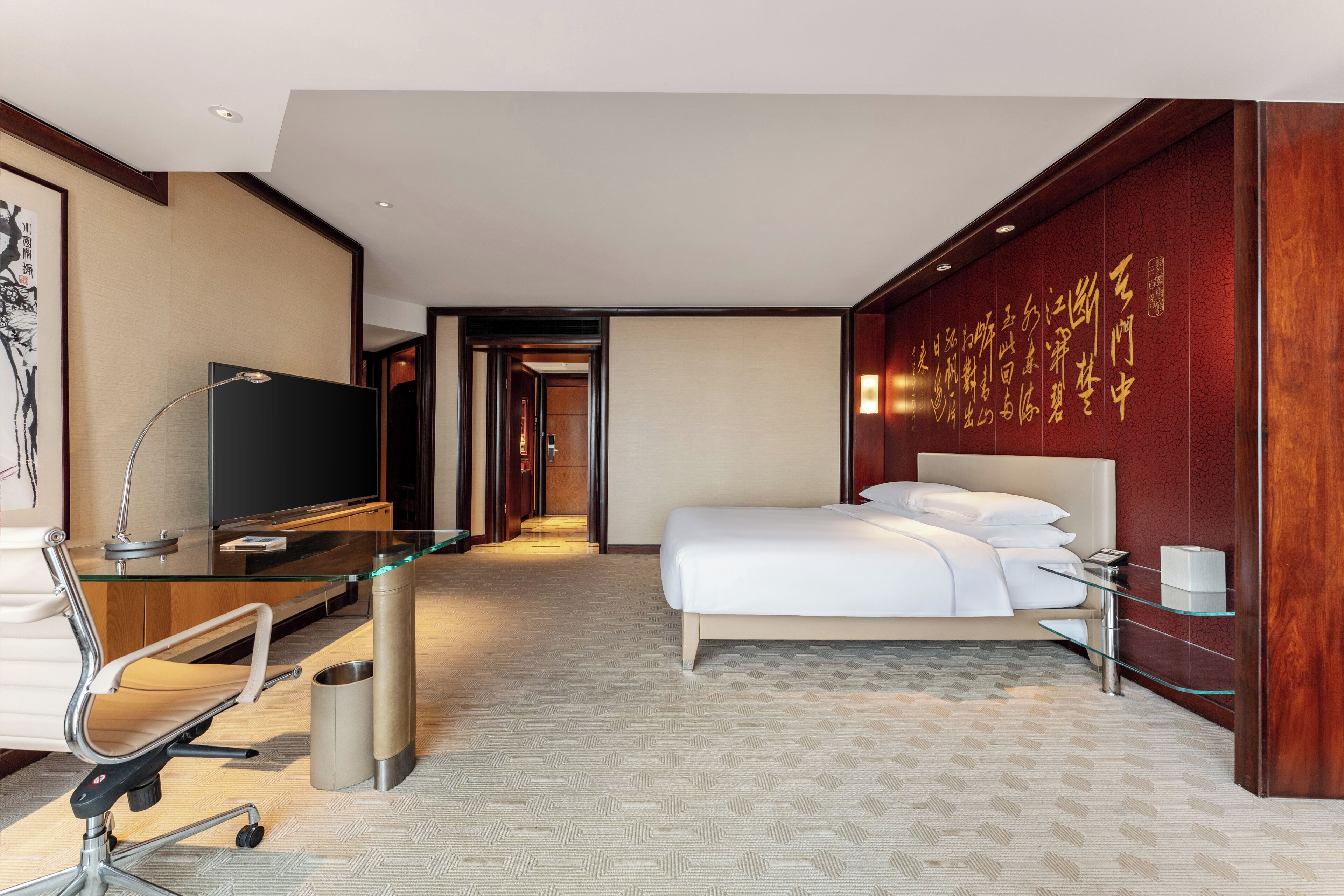 A hotel room with a bed, a desk with a computer, a chair, a TV, and a wall with calligraphy.