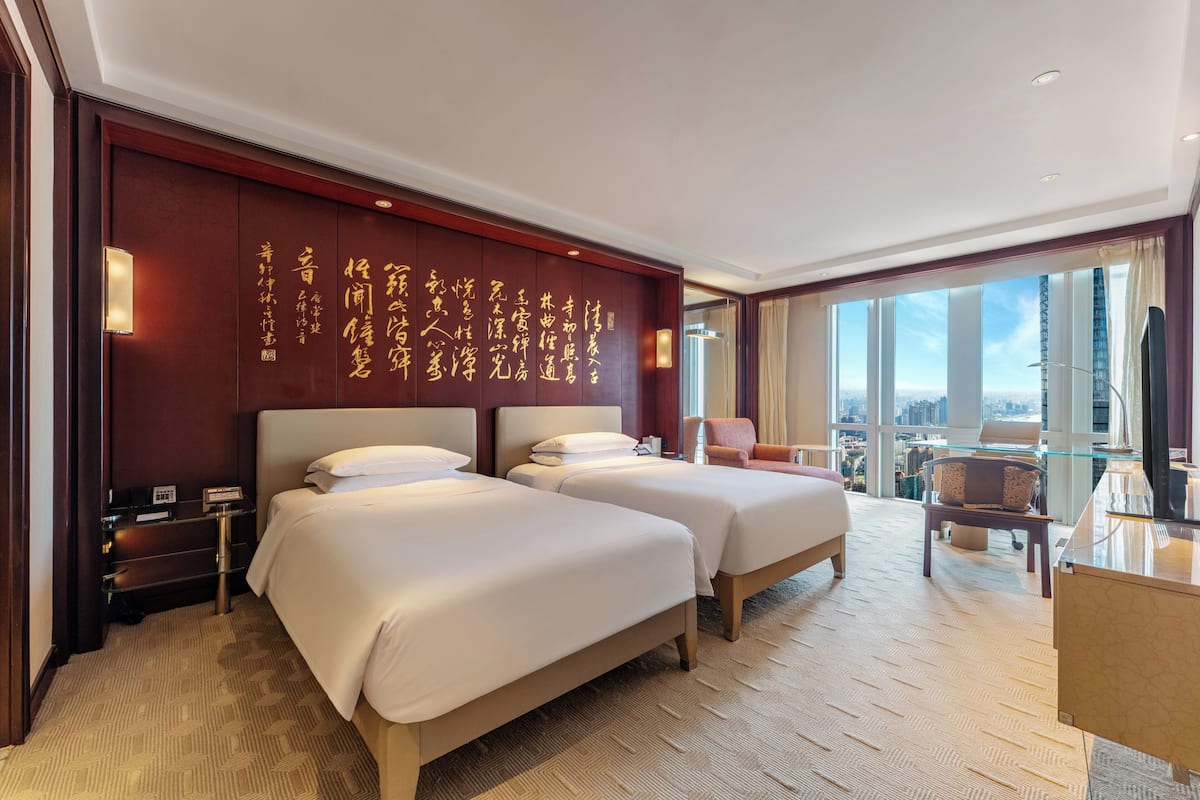 A hotel room with two beds, a desk, a chair, and a large window with a city view.
