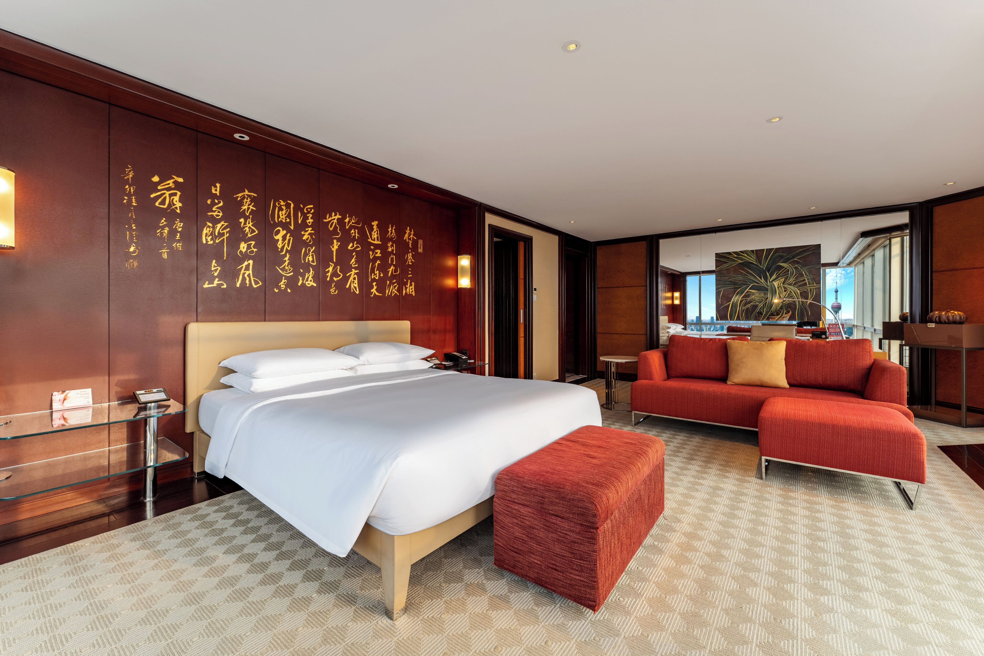 A hotel room with a large bed, a red sofa, a glass side table, and a wall with calligraphy.