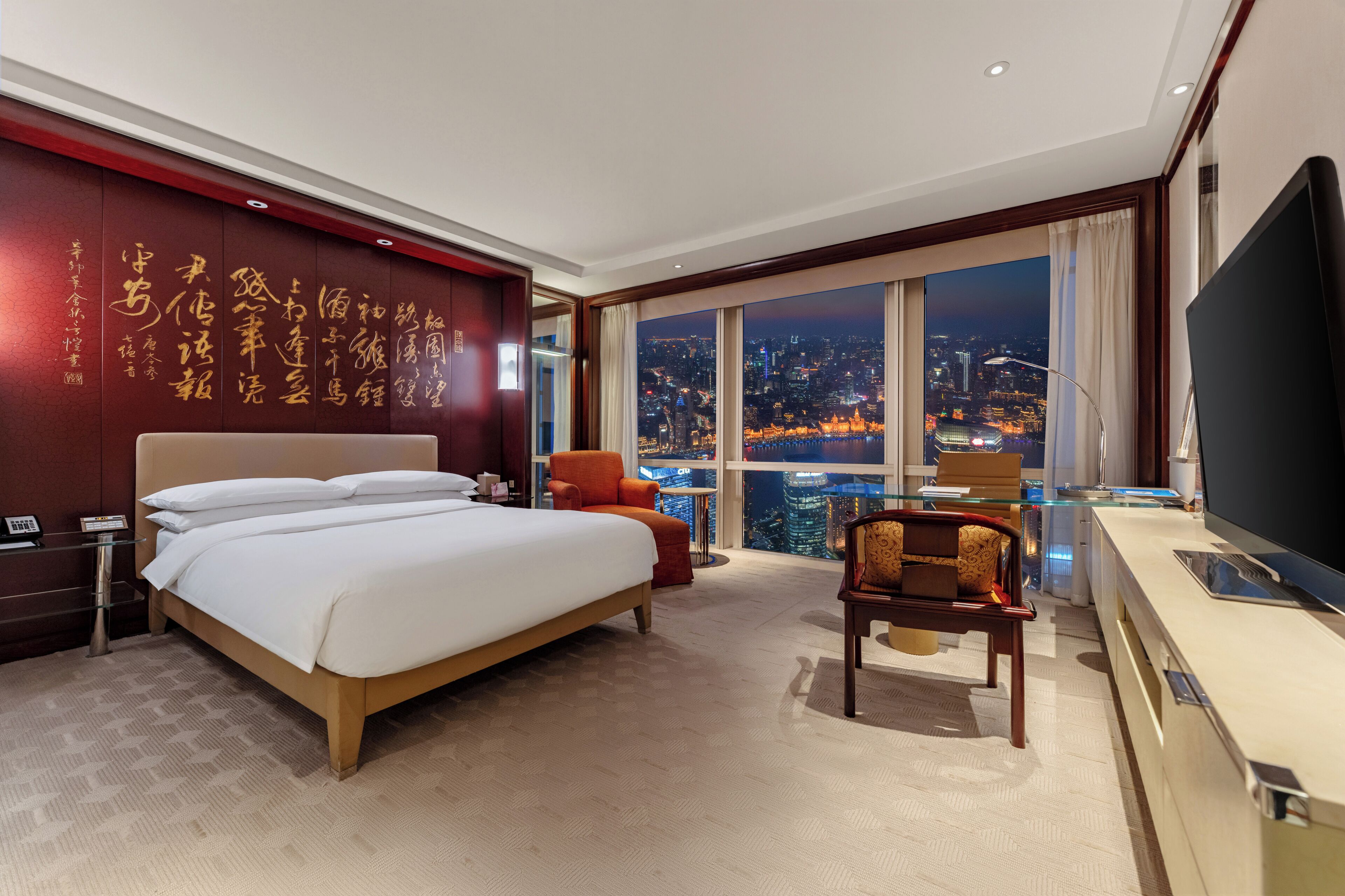 A hotel room with a large bed, a desk with a TV, a chair, and a view of a cityscape at night.