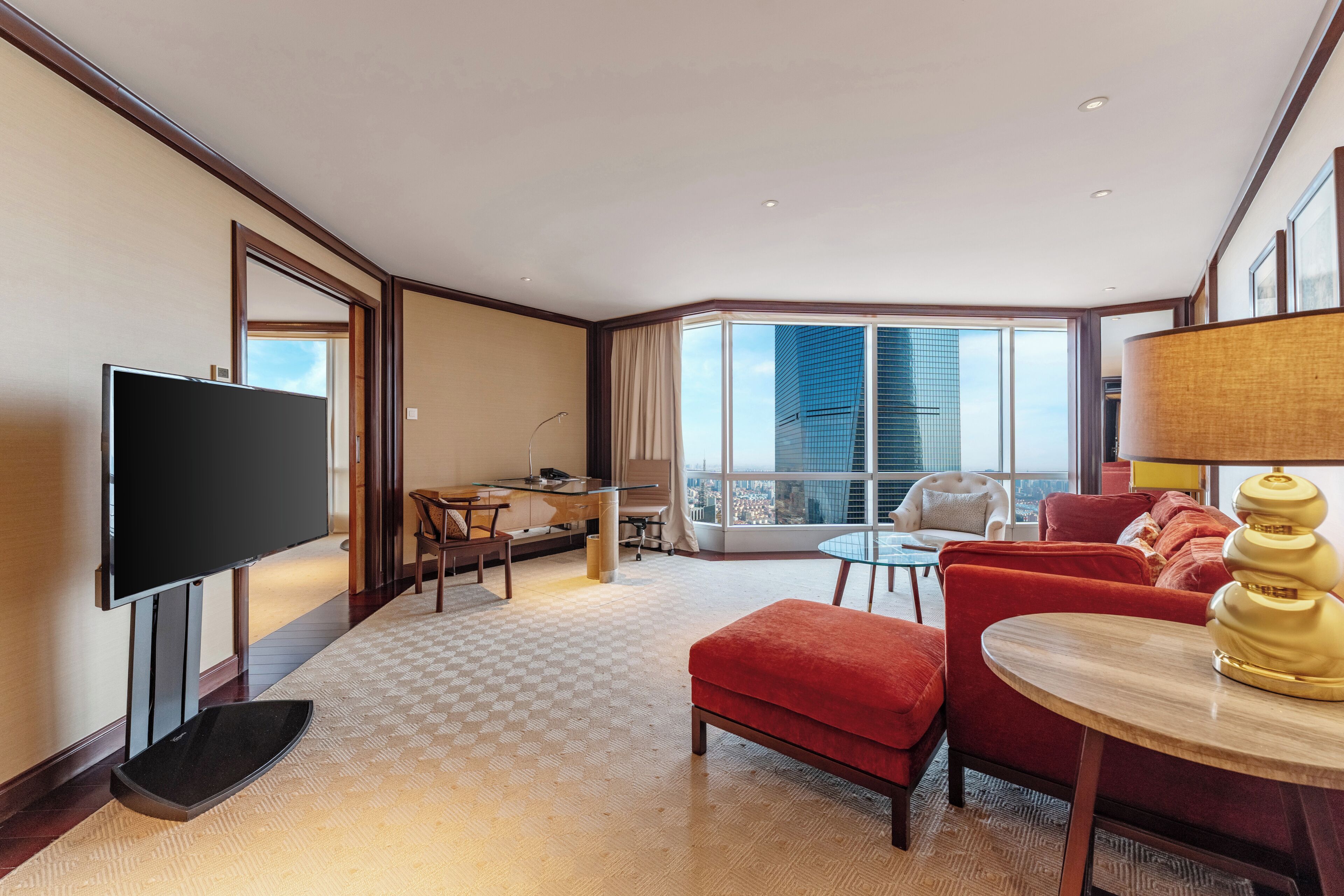 A hotel room with a flat-screen TV, a dining area with a table and chairs, a red sofa, a round coffee table, and a large window offering a city view.
