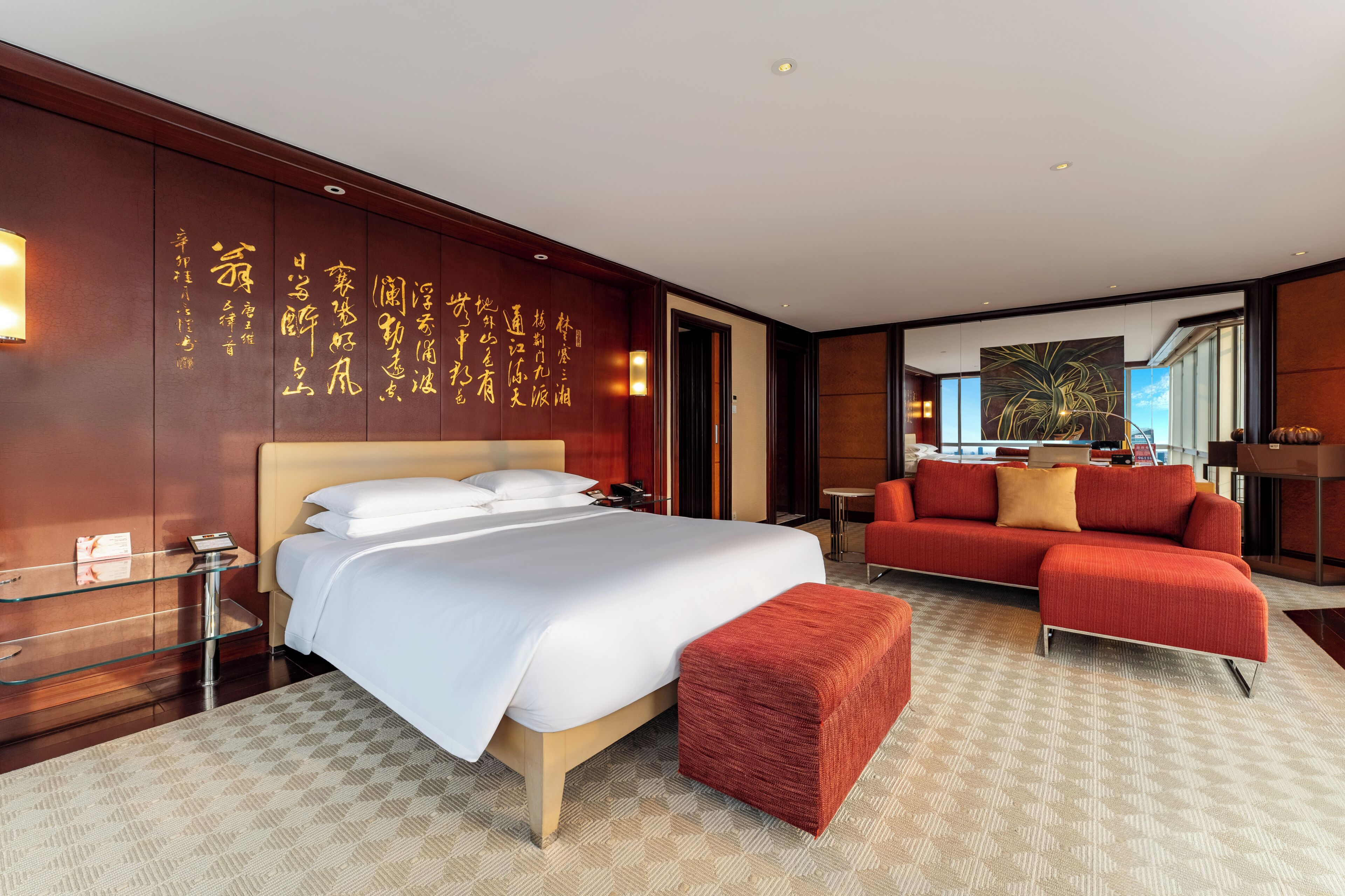 A hotel room with a large bed, a red sofa, a glass side table, and a red ottoman. There is a wall with Chinese characters and a painting on the wall.